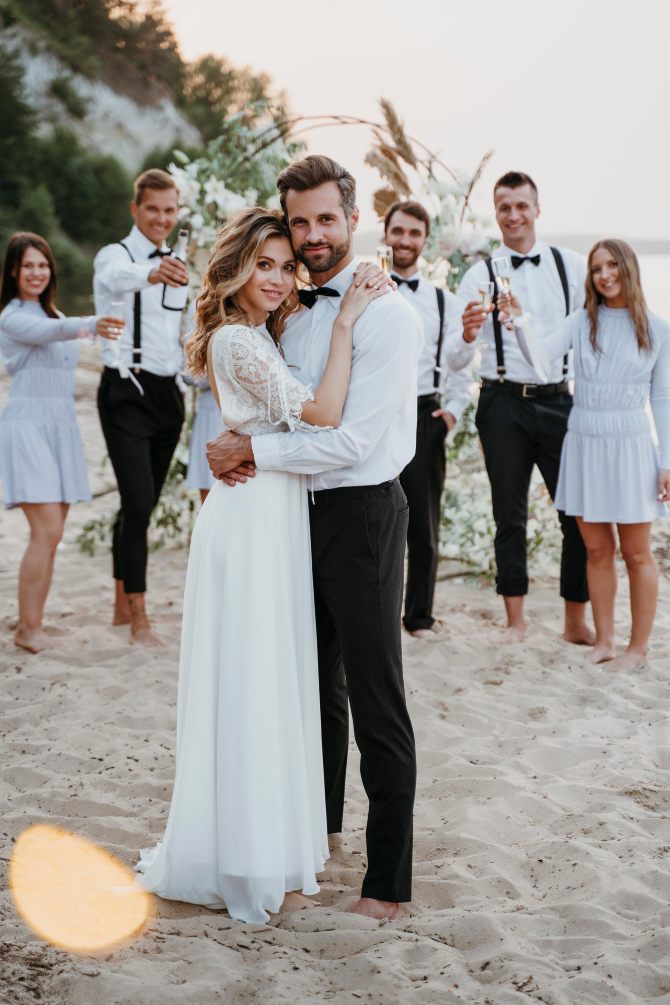 Foto de boda. El novio y la novia se encuentran juntos al frente, y en el fondo están los amigos de ellos. Todos posan para la foto.